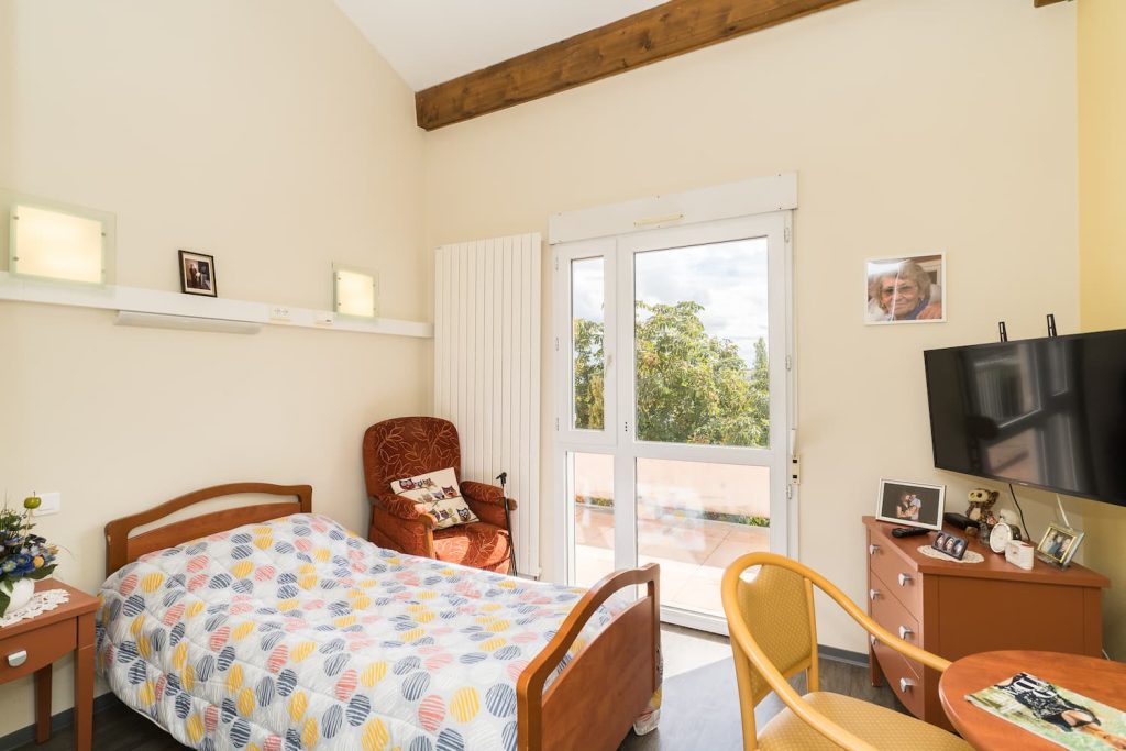 Chambre individuelle avec lit médicalisée, télévision, fauteuil et table, donnant sur le jardin